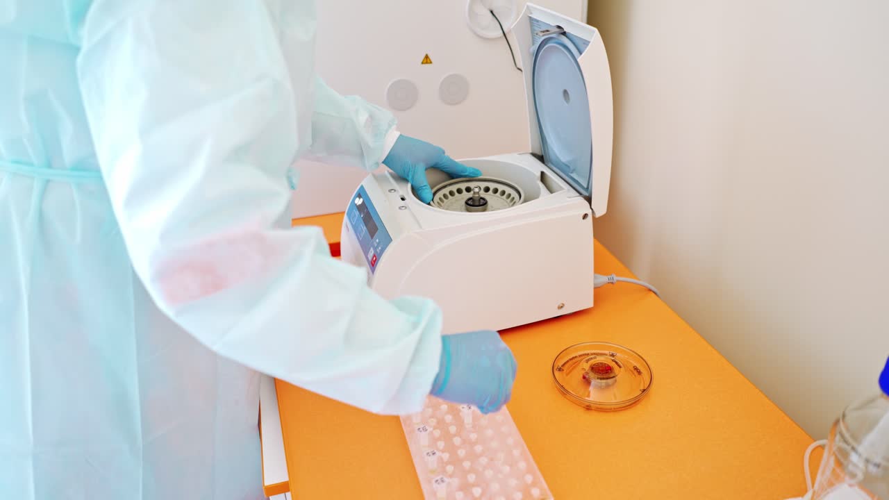 Laboratory assistant near automated device for making analysis. Female worker in protective uniform takes out test tubes from centrifuge in lab.