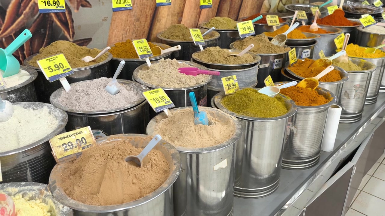 A variety of colorful spices are neatly displayed for sale in clean stainless steel jars. Each container features a clear price tag. The organized market setup looks hygienic and appealing.