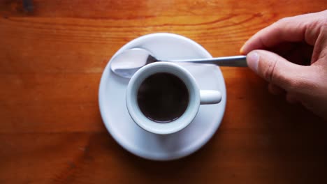 hand is putting spoon next to a hot espresso cup, standing on a brown wooden table