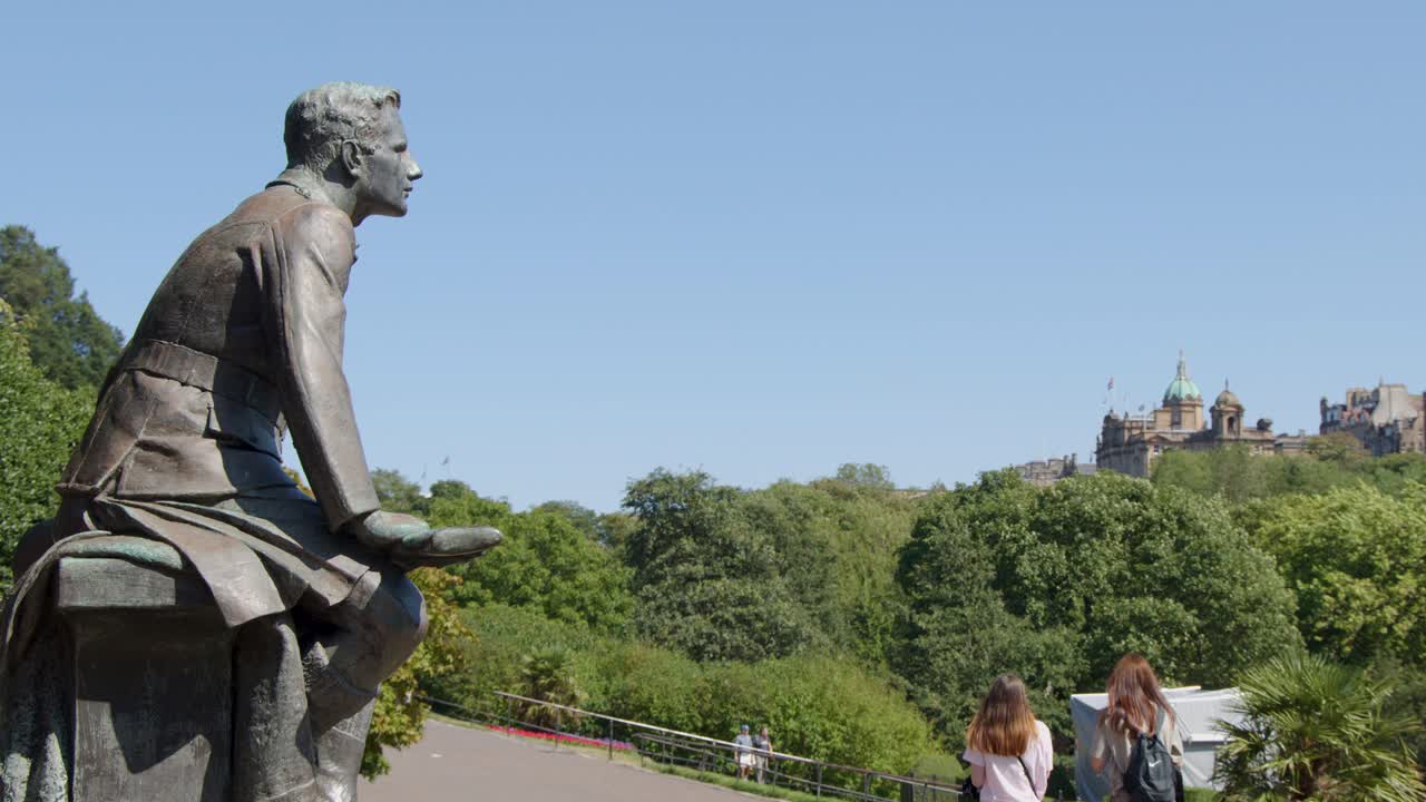 A bronze statue sits on a plinth beside a tree-lined path as people walk by under bright daylight, with historic Edinburgh architecture in the background