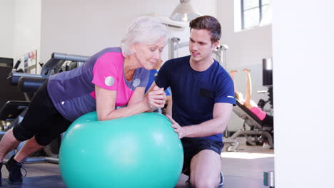 Mujer-Mayor-Haciendo-Ejercicio-Con-Pelota-Suiza-Siendo-Alentada-Por-Un-Entrenador-Personal-En-El-Gimnasio