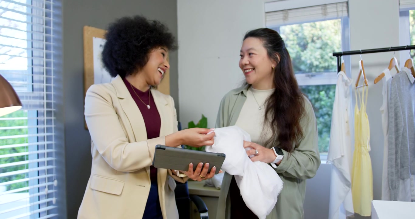 Female coworkers, consultant tapping tablet and stylist touching garment, selecting at showroom