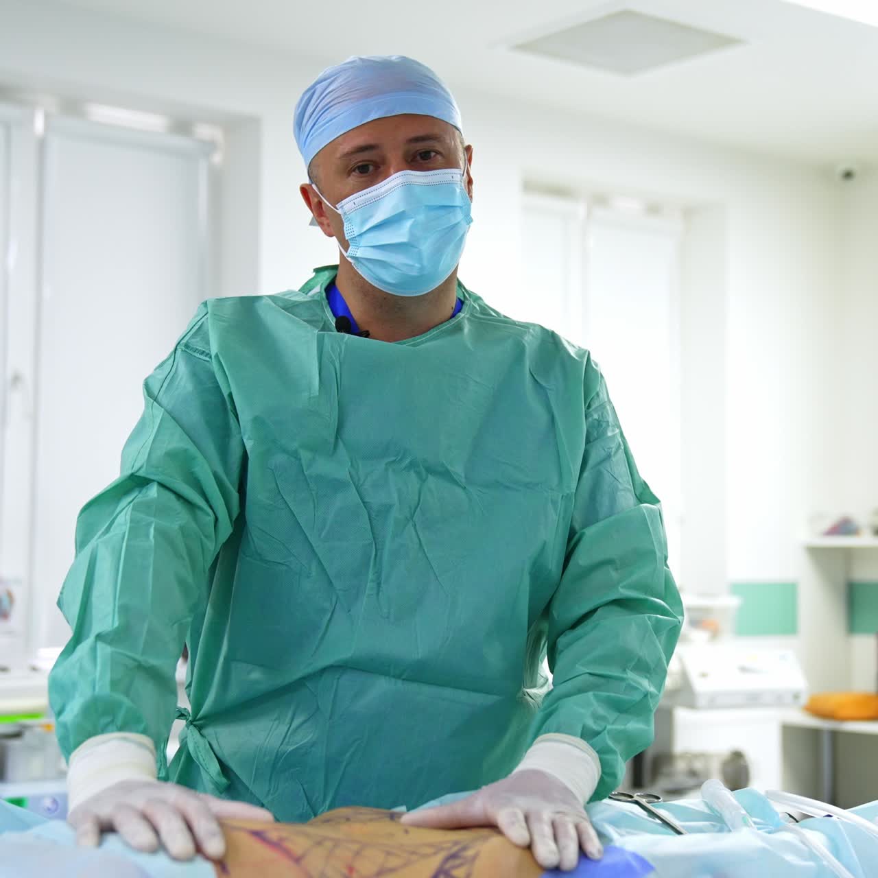 Male doctor in cap and mask stands over the patient prepared for liposuction surgery. Experienced surgeon talks to camera pointing at and touching the female patient belly