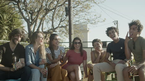 happy friends looking at camera, making funny faces and laughing while sitting on rooftop during an outdoor party