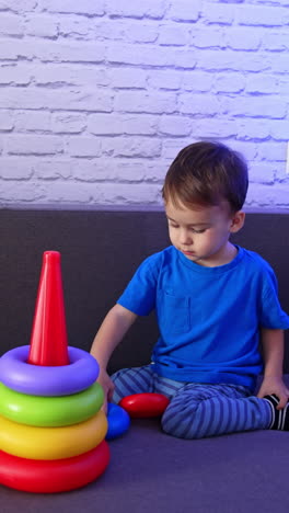 calm little baby boy wearing blue t-shirt playing with toy sitting on the sofa. cute toddler assembling the pyramid. vertical video