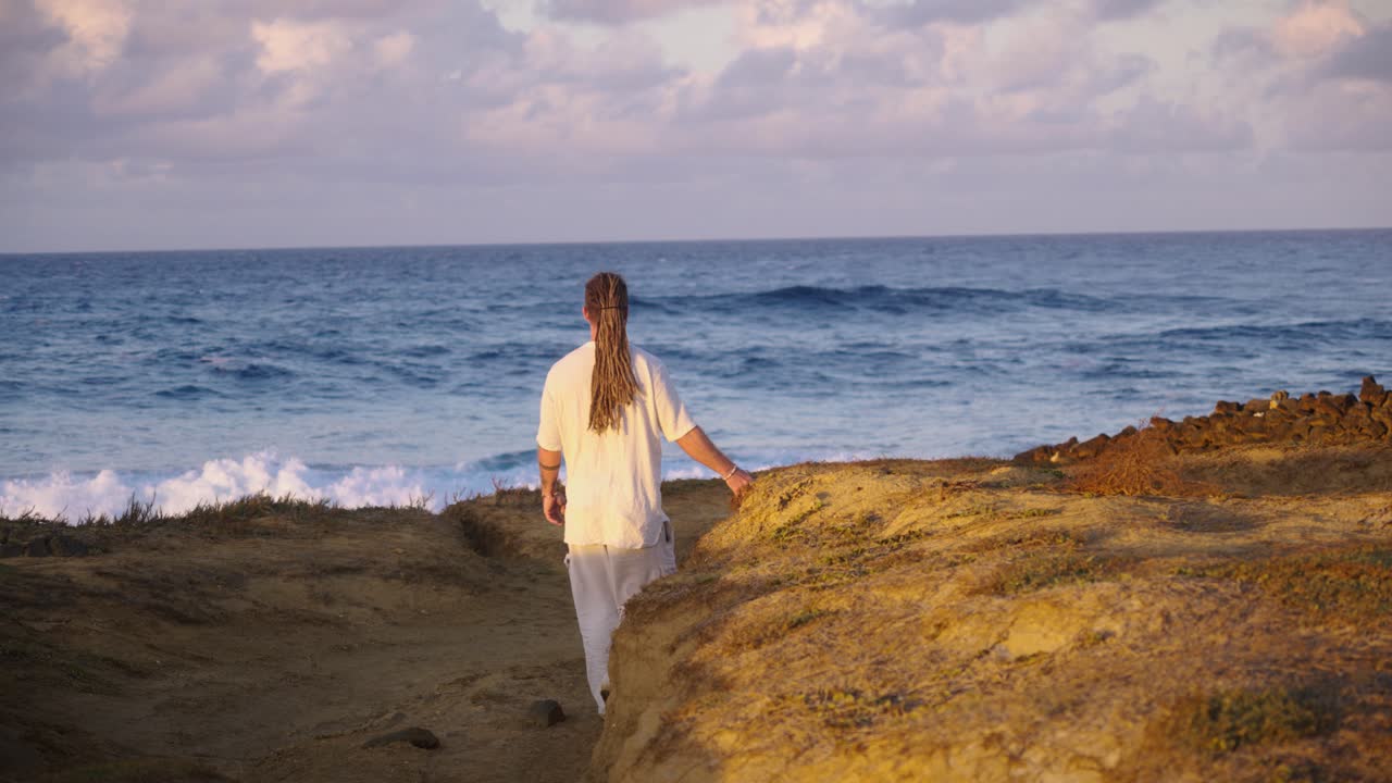 A man in white clothing walks away from the camera with his arm outstretched, moving toward crashing waves and the open ocean horizon in daylight