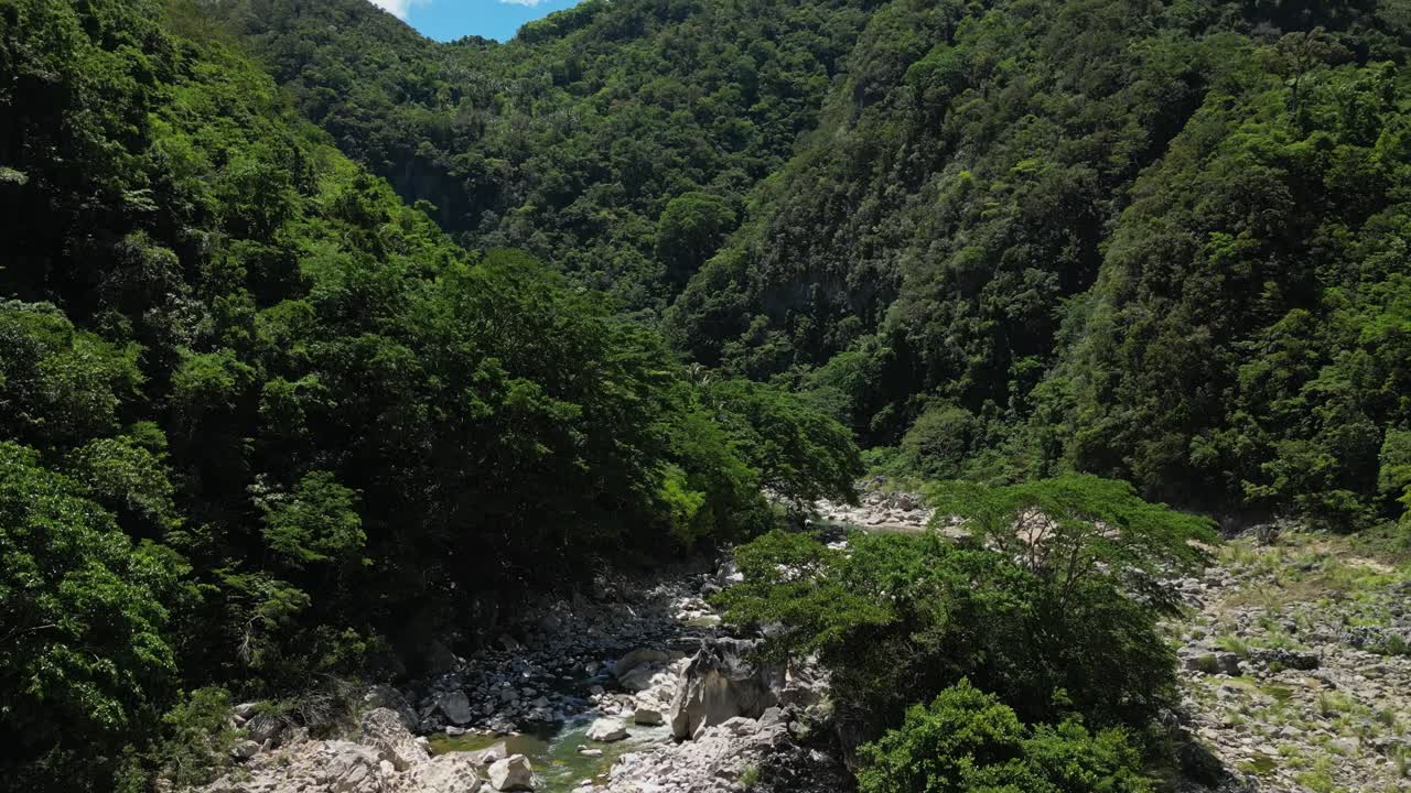Shot from below rising upward showing river, rocks with plants, then shifting to top view of lush green mountains around Tinipak River, Philippines