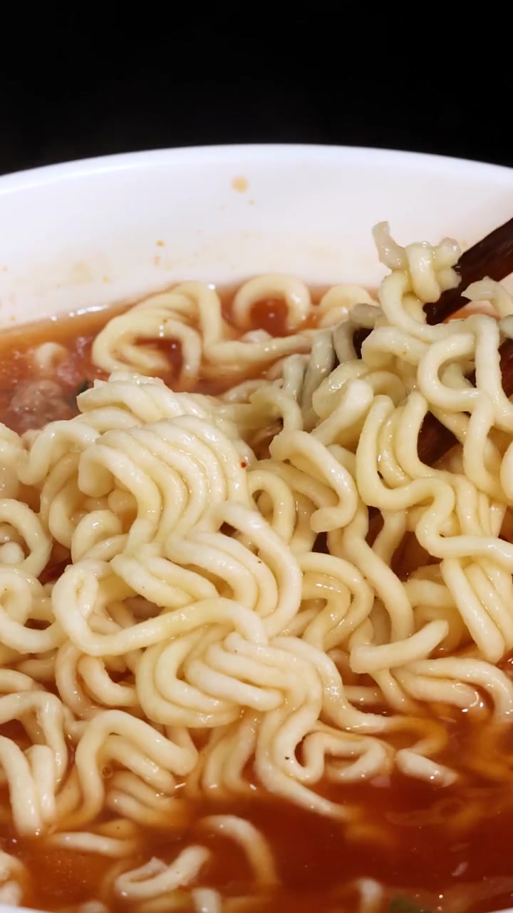 Sepasang sumpit mengangkat mi instan yang sudah matang dari mangkuk mengepul berisi kuah merah di bawah pencahayaan terang dan merata. Kamera close-up statis menangkap tekstur dan gerakan.