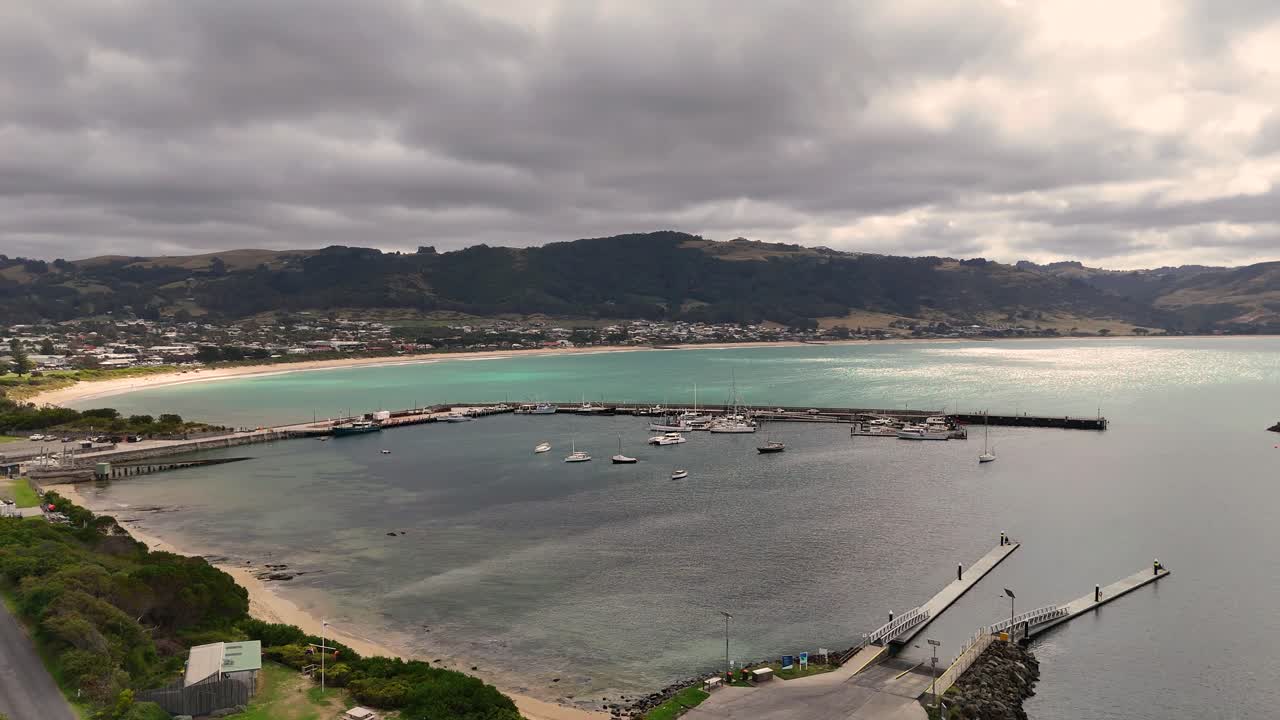 Drone footage captures Apollo Bay's serene harbour and coastline under cloudy skies, showcasing boats, piers, and lush landscapes