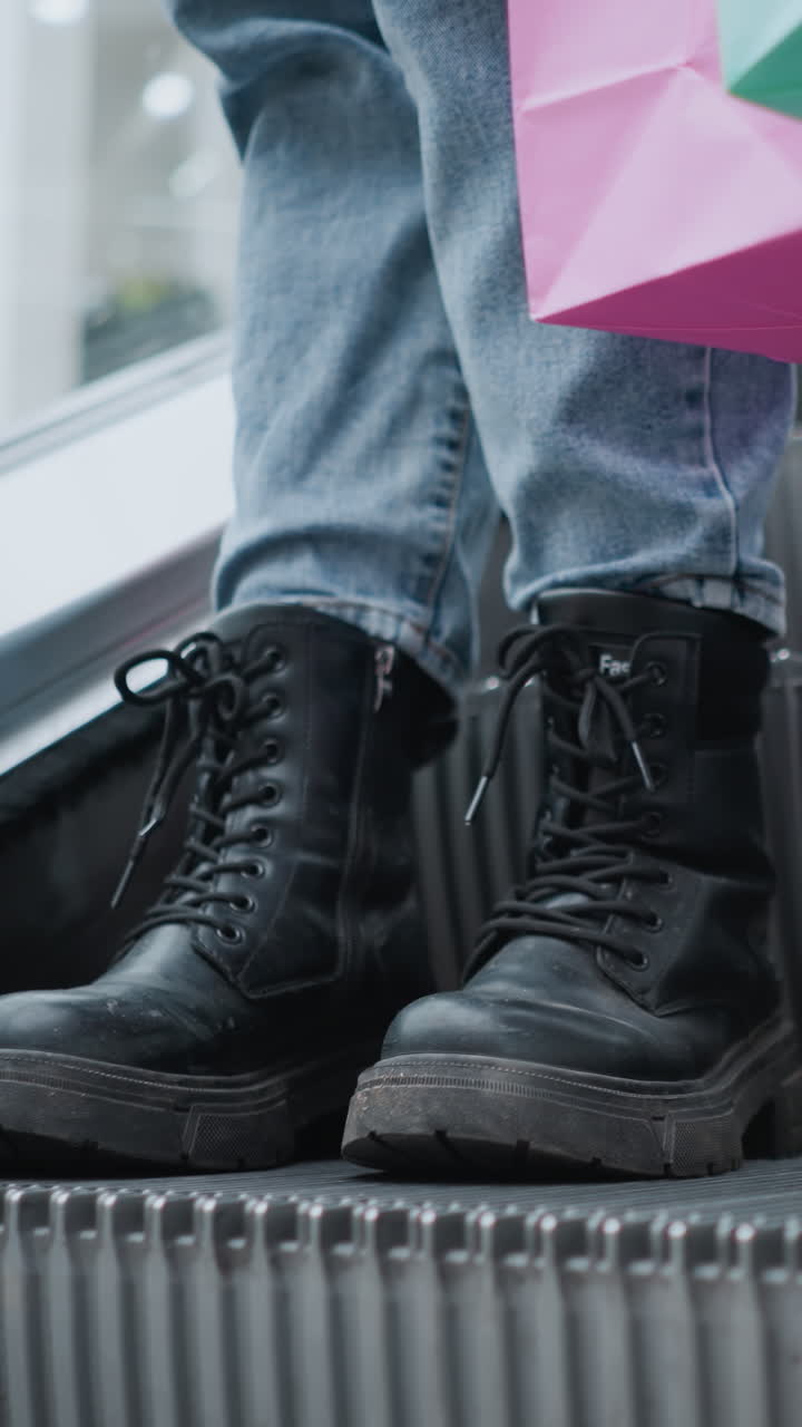 primer plano de piernas en botas negras y vaqueros de pie cerca de la barandilla de la escalera mecánica mientras sostiene bolsas de compras en un vibrante centro comercial, la escena captura una elegante experiencia de compras urbanas con detalles modernos