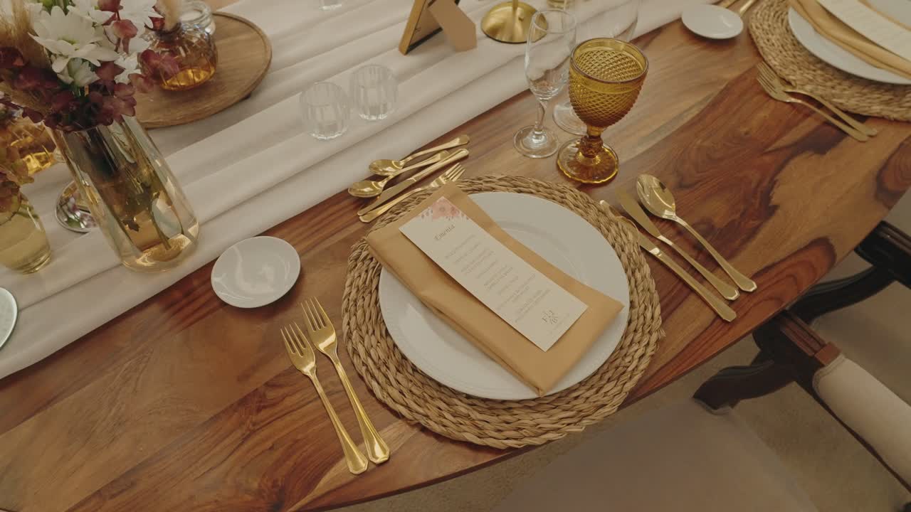 Vista dall'alto di una raffinata disposizione della tavola con piatto bianco, tovaglietta intrecciata, posate dorate, menu e decorazioni floreali.