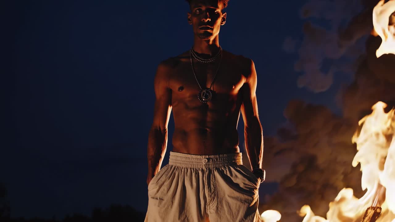 Male figure stands confidently in front of a roaring fire, showcasing strength and intensity, with dark night sky creating a dramatic backdrop
