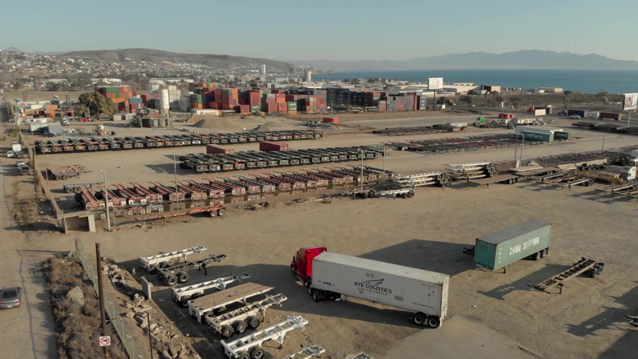 Aerial drone view of industrial container yard with trucks and cargo storage area. Shipping containers, trailer chassis and freight equipment fill expansive logistics facility near coastline