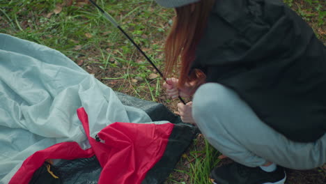 aerial view shows woman squatting and fixing flexible tent pole into ring of tent fabric while hooking fabric clip around it, scene captures precise motion on forest ground with gear visible