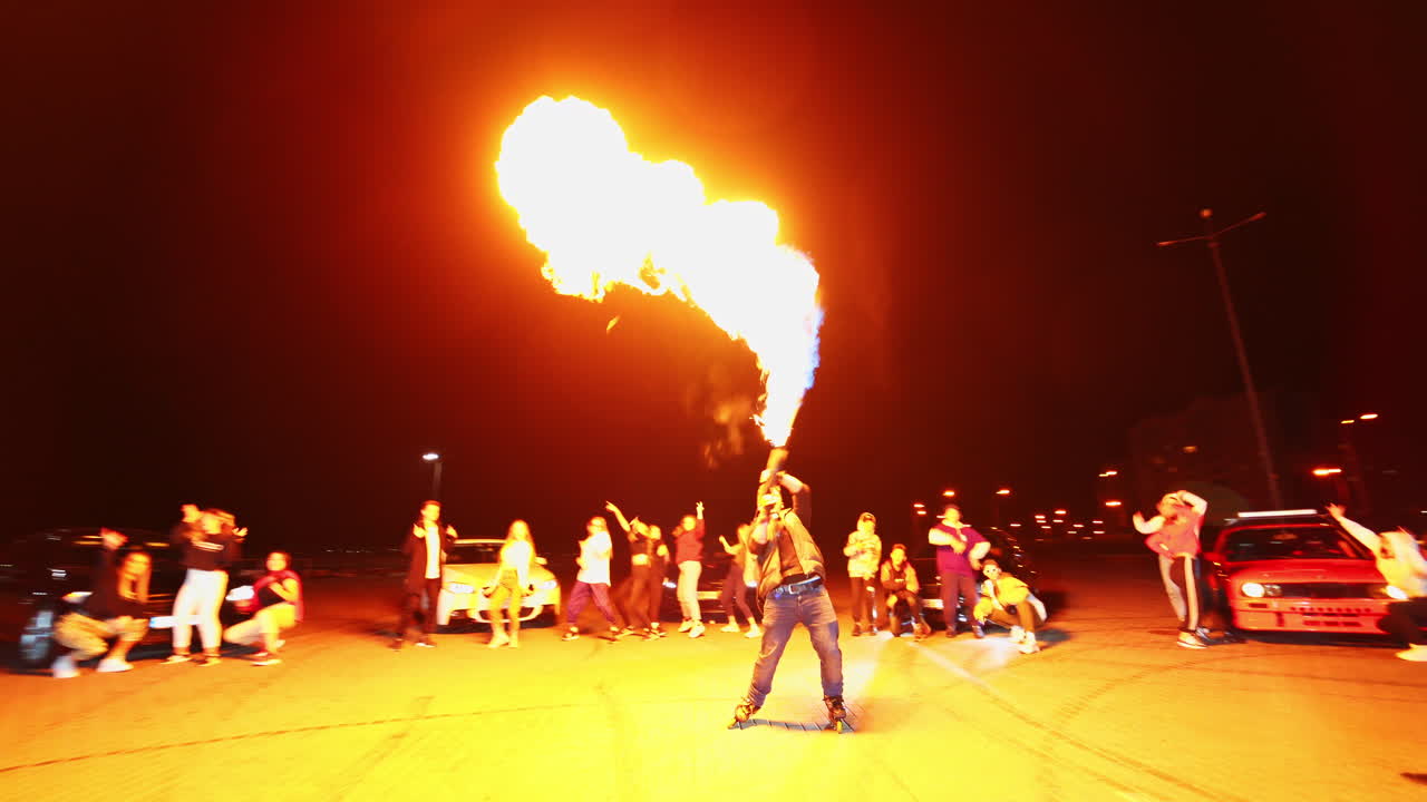 Caucasian man shooting fire from a special gun outdoors at night. Young people dancing standing near race cars at backdrop.
