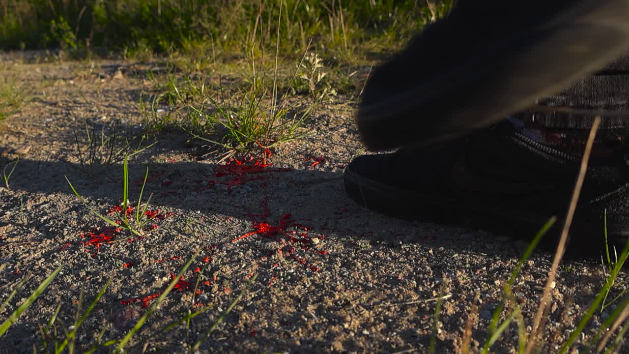Red blood falling on a sandy and muddy road outdoors in sunny day at slow motion and a white diverse hand is placing a yellow police crime marker next to it, forensic investigation scene, bokeh