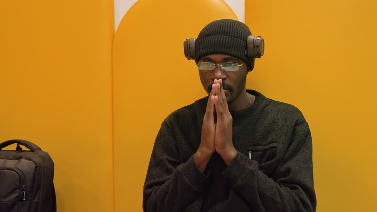 Black man praying at desk in yellow acoustic booth, wearing beanie, headphones and glasses, hands clasped beside laptop and backpack contemplative pause between podcast takes and study session