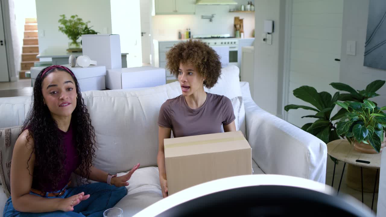 Donne afroamericane sedute sul divano in soggiorno che tengono una scatola e filmano un unboxing con un ring light.