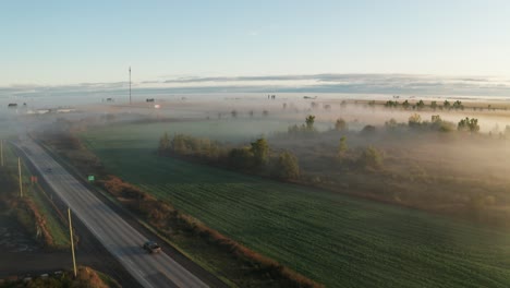 trucks and cars on road driving through farmland early in morning with fog covering fields and hanging in trees