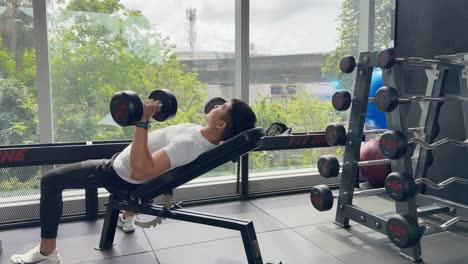 a man exercises with dumbbells on an incline bench in a well-lit gym, surrounded by weights and large windows
