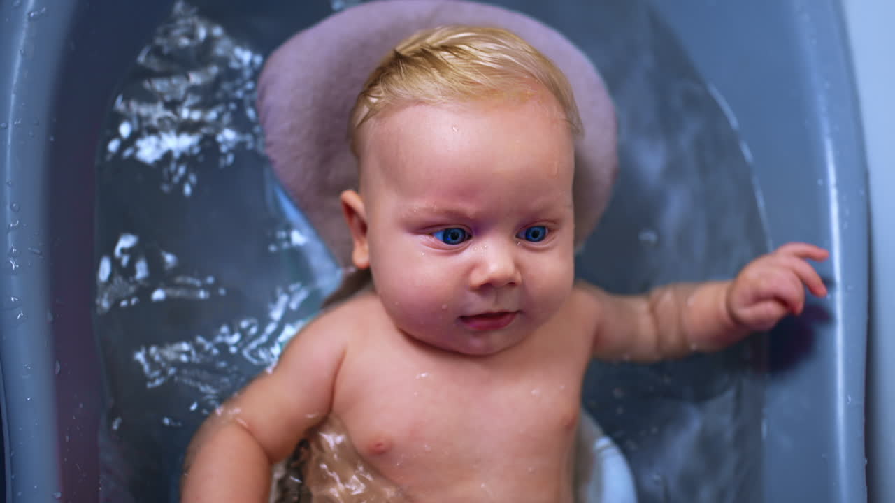 Sweet blue-eyed baby boy with fair hair lies in the wash tub. Happy kid waving his hands and feet while having a bath. Close up.