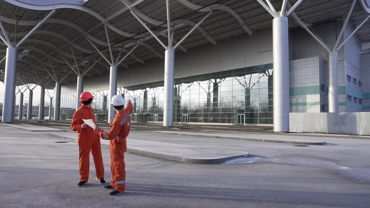 trabajadores de la construcción en uniforme naranja y cascos duros mirando los planes juntos. edificio en el fondo