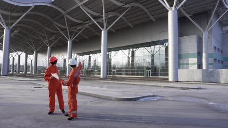 Bauarbeiter-In-Orangefarbenen-Uniformen-Und-Helmen-Schauen-Sich-Gemeinsam-Die-Pläne-An.-Gebäude-Im-Hintergrund