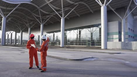 construction workers in orange uniform and hardhats looking over plans together. building at the background