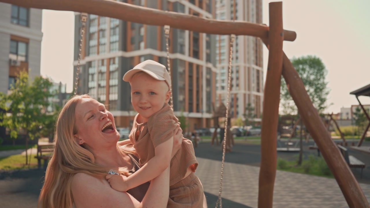 Frau und kleiner Junge verbringen gemeinsam Zeit auf dem Spielplatz, wobei die Mutter ihr Kind auf der Schaukel anschiebt. Die Szene zeigt Elemente eines Outdoor-Parks wie Schaukeln und Bänke vor einer urbanen Kulisse hoher Gebäude.
