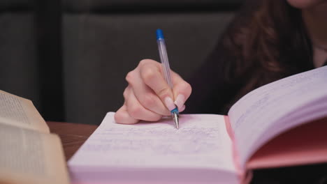 close up of female hand holding pen writing in pink notebook filled with text while brown book lies open on wooden table, creating focused workspace scene filled with detail and calm ambiance