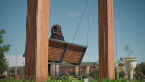 woman on swing chair swinging with hair fluttering in the wind, trees in the background, and a blurred person in the distance in a public park