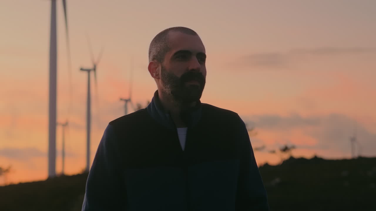 A cheerful Caucasian young man with short hair and blue jacket enjoys sunset near wind turbines in Portugal. Cinematic shot symbolizes renewable energy and sustainability, showcasing nature's beauty.