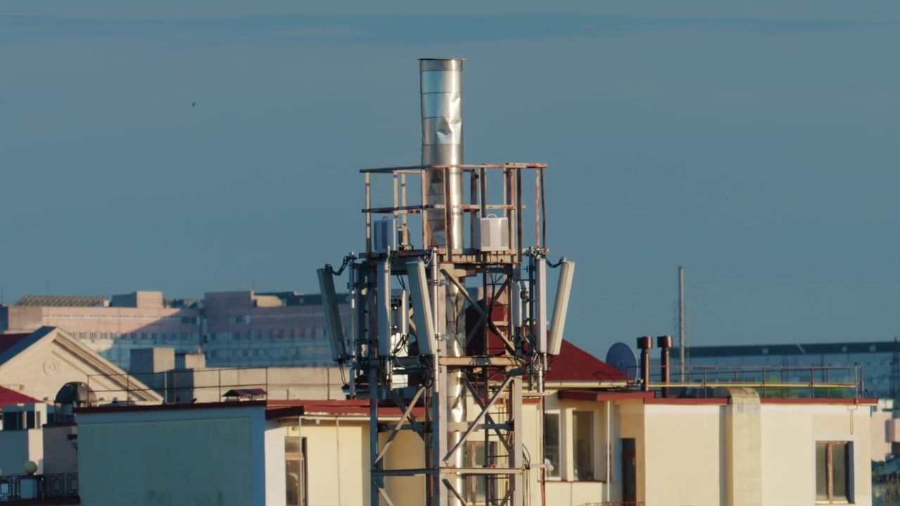 Urban rooftop scene featuring a cellular communication tower with multiple panel antennas, mounted on a metal framework above residential buildings