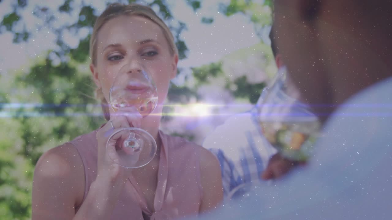 Woman raising glass toward offscreen-guest tasting wine in yard while flare moving on glass for ads