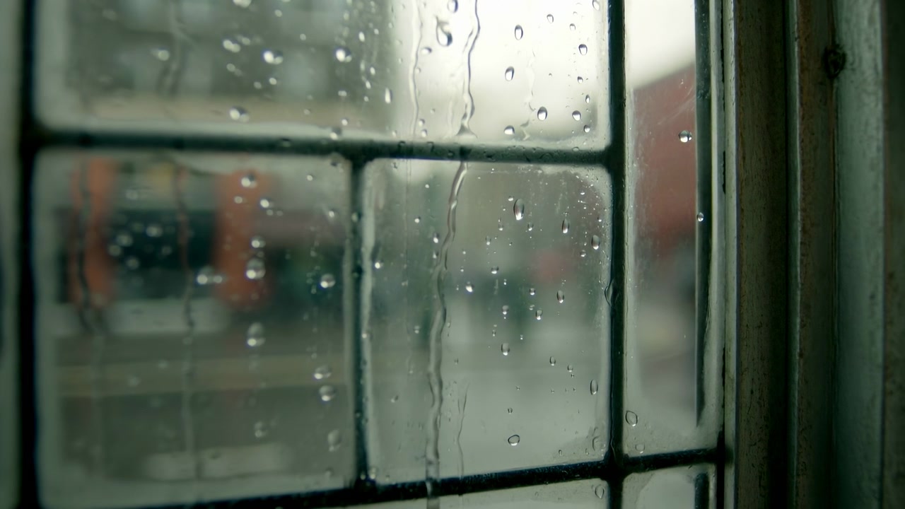 Close up of an old leaded glass window with raindrops and vertical streaks on textured panes. Soft overcast light falls across a metal frame and sill at the