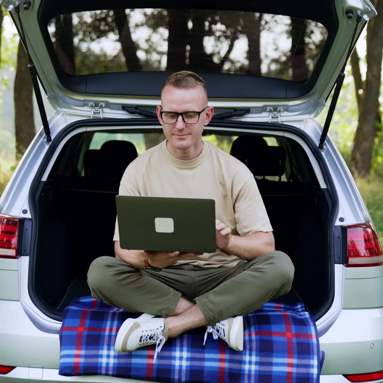 Caucasian man sitting in the trunk of his car puts on glasses. Freelancer is getting ready to work on his laptop. Beautiful summer nature at backdrop