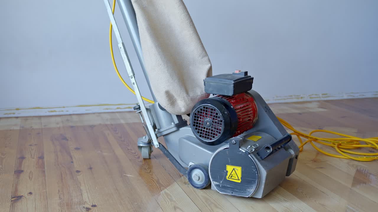 Electric floor sander stands on partially sanded pine wood floor, dust collection bag and power cable attached, positioned indoors against a plain wall, used for professional parquet restoration work.