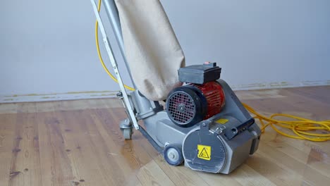 electric floor sander stands on partially sanded pine wood floor, dust collection bag and power cable attached, positioned indoors against a plain wall, used for professional parquet restoration work.