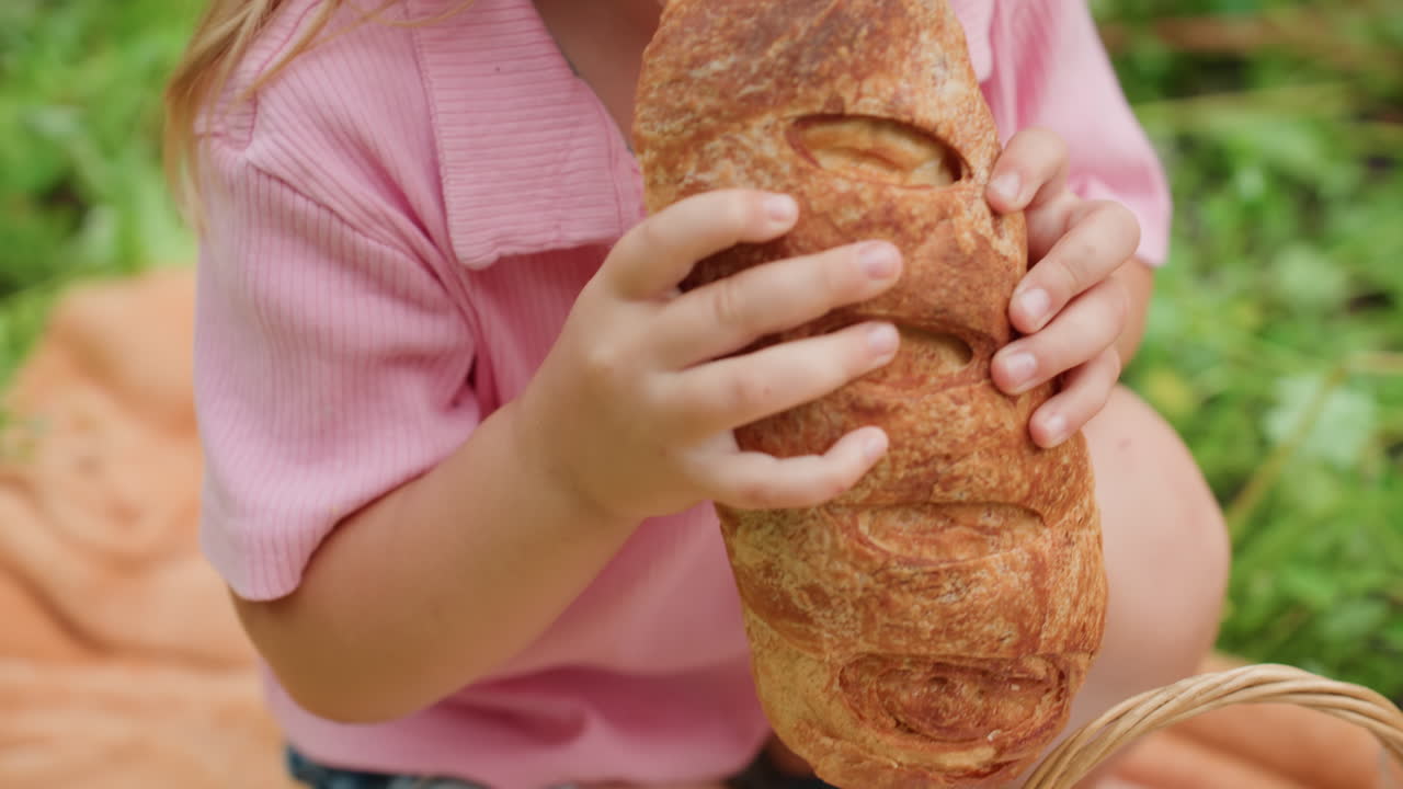 niño blanco sujetando una hogaza grande sobre una manta de pícnic, agarrando el pan con sus pequeñas manos, levantando la hogaza para dar el primer bocado grande, primer plano de la corteza de sésamo y los dedos, expresión concentrada, textura crujiente, verano