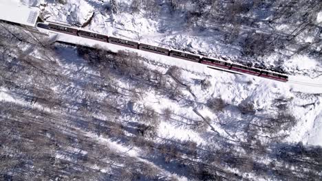 Aerial-reveal-of-the-Bernina-Express-panorama-train-going-through-a-snow-covered-mountain-winter-landscape-on-a-sunny-day-in-Alp-Grum,-Switzerland