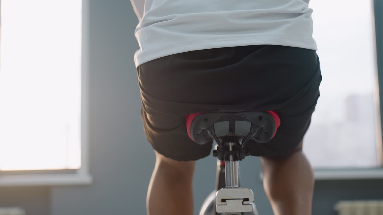 back view of man in white top pedaling stationary bike in sunlit gym, muscles engaged, urban skyline blur outside window, focused exercise session with modern equipment and bright ambient light