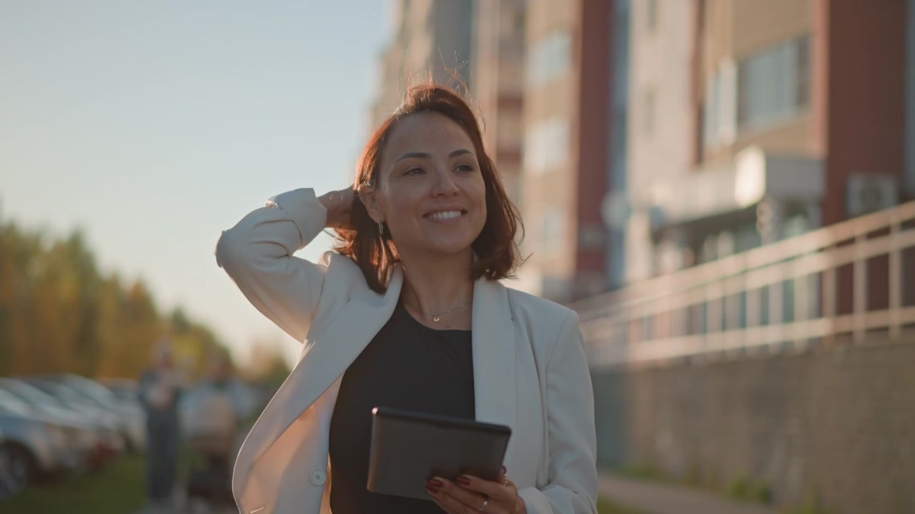 Mulher de blazer claro segura e interage com um tablet enquanto caminha ou faz uma pausa em uma passarela urbana; o movimento visível dos dedos e uma leve inclinação para frente indicam uma ação de tocar ou rolar com propósito.