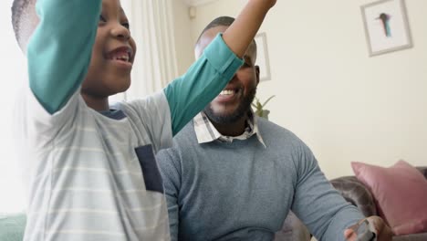 happy african american father and son sitting on sofa using tablet and high fiving, in slow motion