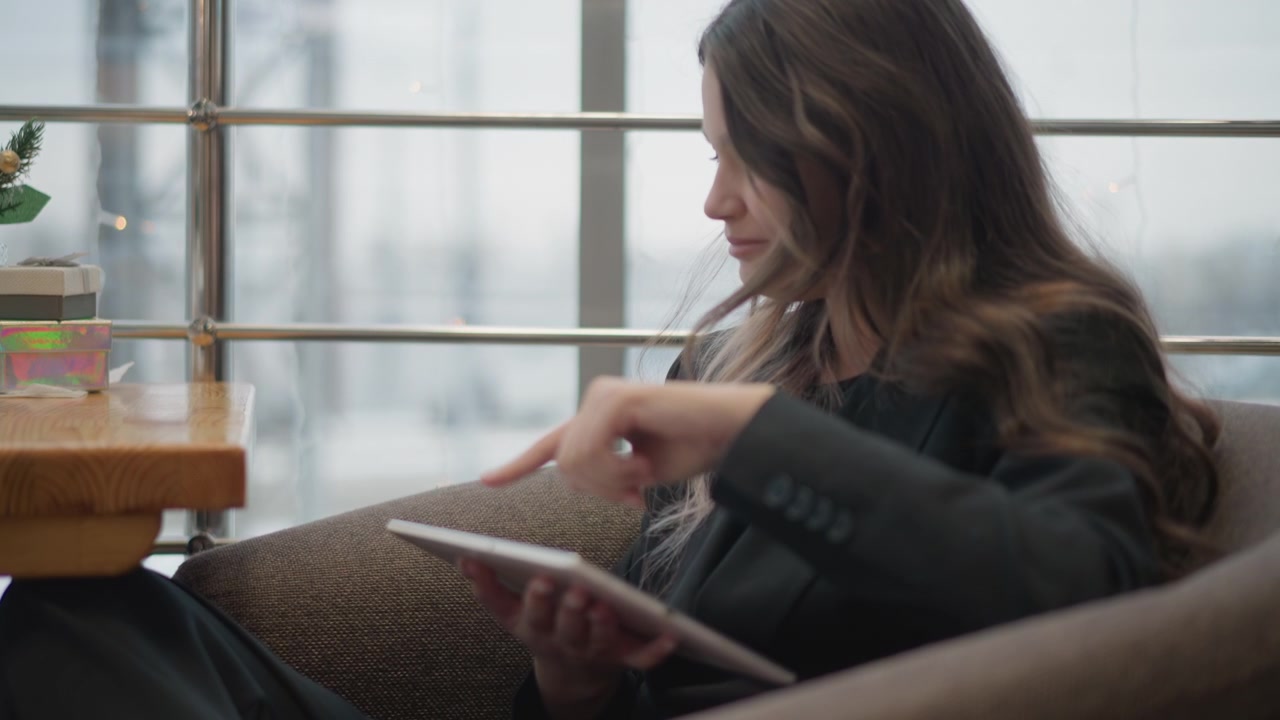 Smilende ung professionel kvinde i en polstret lænestol, med krydsede ben og lænet frem, mens hun tapper på en tablet med pegefingeren, bruger en rolig caféindstilling til afslappet digitalt fokus og en uformel stemning.