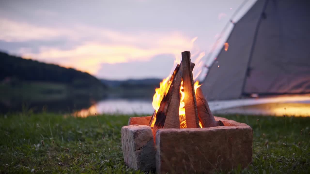 スロベニアのサヴチで、夕暮れ時にテントと穏やかな湖の近くの草地に明るい炎が燃えるキャンプファイヤー。平和なアウトドアキャンプの雰囲気と自然の風景を捉えている。