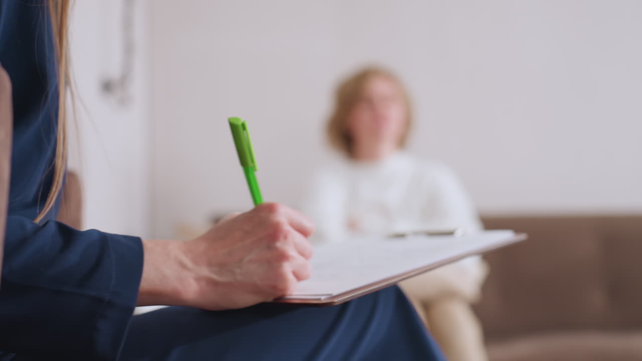Counselor holding green pen and writing on clipboard while seated in conversation with client, who appears blurred in background, showing attentive listening and documentation during therapy session