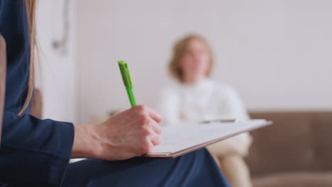 counselor holding green pen and writing on clipboard while seated in conversation with client, who appears blurred in background, showing attentive listening and documentation during therapy session