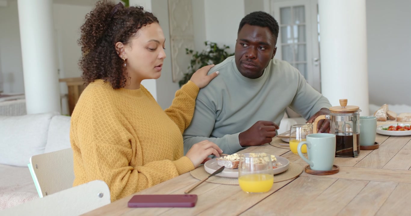 African couple eating meal at home, man eating pastry, woman touching him, using purple phone
