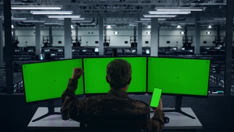 vista trasera de los militares asiáticos felices mirando la pantalla verde del teléfono móvil y celebrando mientras trabajan con múltiples monitores de computadora en el centro de datos