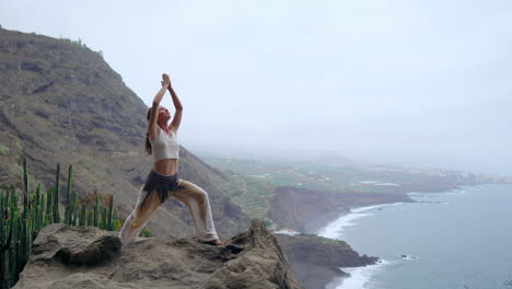 As-the-sun-sets-over-the-blue-ocean,-a-young-woman-performs-yoga-on-a-rocky-seashore,-representing-a-healthy-lifestyle,-harmony,-and-the-interconnectedness-of-humans-and-nature